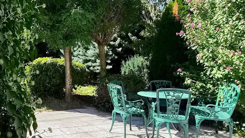 Gepflasterte Terrasse mit grünen Gartenmöbeln, umgeben von üppiger Bepflanzung und Bäumen.