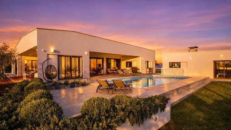 Moderne Villa mit beleuchtetem Poolbereich und Garten bei Sonnenuntergang, ideal für entspannte Abende.