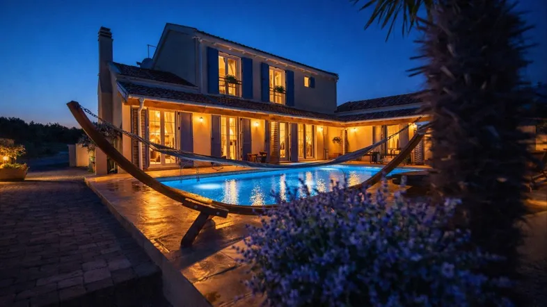 Beleuchteter Außenbereich mit großem Pool, Hängematte und mediterraner Architektur bei Nacht.