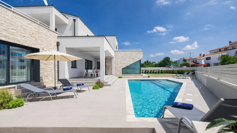 Moderne Villa mit großem Poolbereich, Sonnenliegen und überdachter Terrasse unter blauem Himmel.