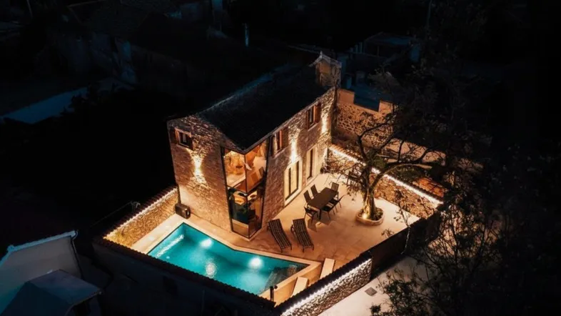Nachtaufnahme eines Hauses mit beleuchtetem Poolbereich und möblierter Terrasse unter sternenklarem Himmel.