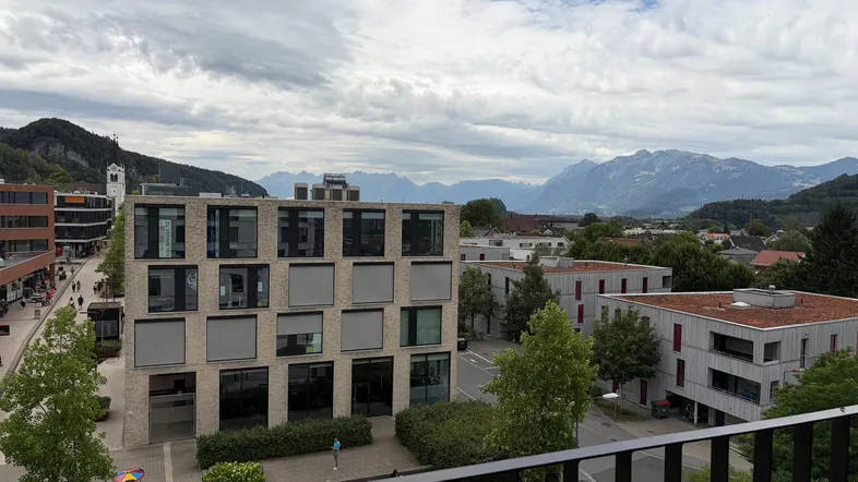 Blick auf moderne Gebäude, grüne Hügel und eine beeindruckende Bergkette unter bewölktem Himmel.