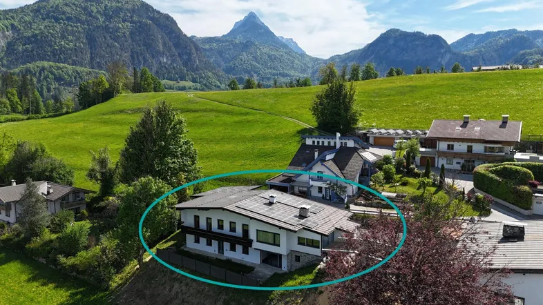 Luftaufnahme eines Hauses in alpiner Landschaft mit grünen Hügeln und Bergen im Hintergrund.