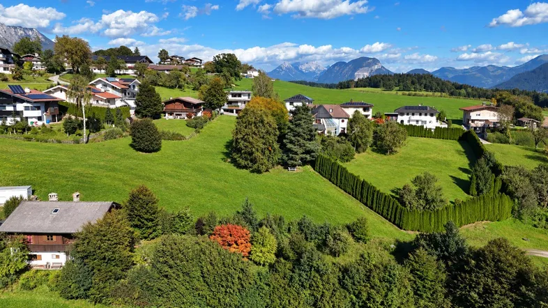 Panoramablick auf eine ländliche Wohnsiedlung mit grünen Hügeln und majestätischen Bergen im Hintergrund.
