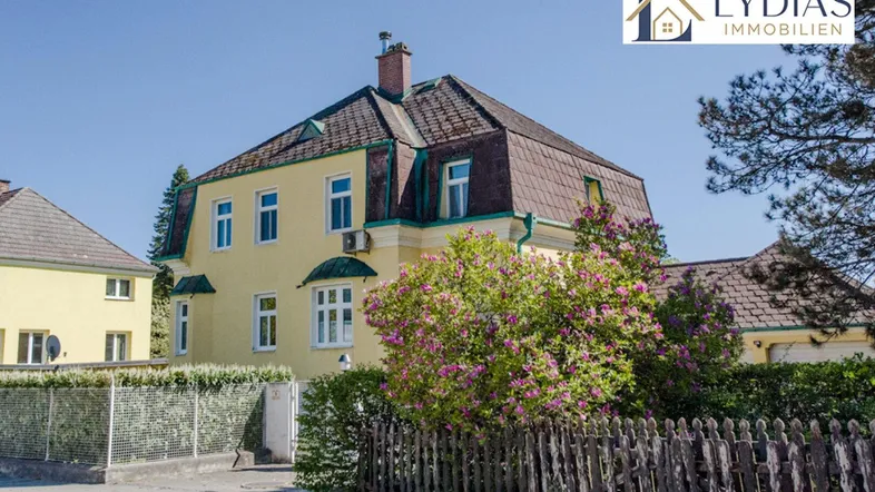 Historische Stadtvilla mit markantem Mansarddach, umgeben von einem Holzzaun und blühenden Büschen.