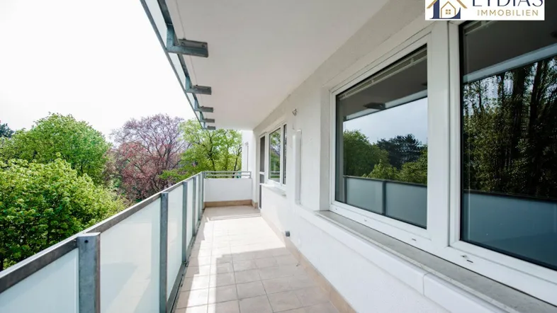 Langer, gefliester Balkon mit Glasgeländer und Blick ins Grüne, ideal zum Entspannen im Freien.