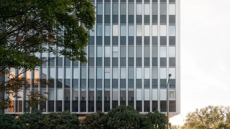 Nahaufnahme der modernen Gebäudefassade mit großen Fenstern und grünen Bäumen im Vordergrund.
