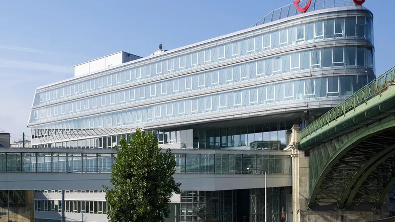 Modernes Bürogebäude mit geschwungener Glasfassade und angrenzender Brückenkonstruktion unter blauem Himmel.