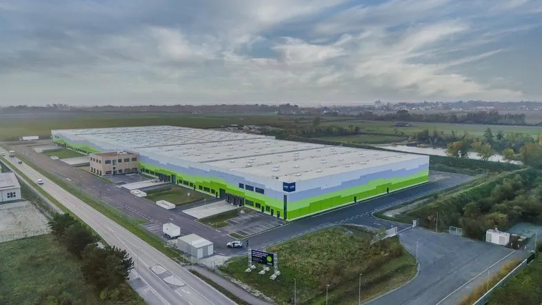 Luftaufnahme eines großen modernen Logistikzentrums mit grünen und blauen Fassaden unter bewölktem Himmel.
