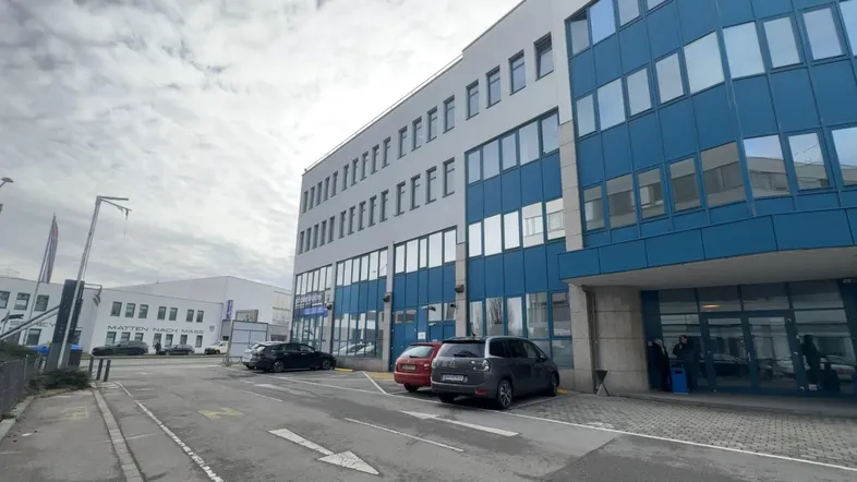 Modernes Bürogebäude mit blauer Glasfassade und Parkplätzen davor unter bewölktem Himmel.