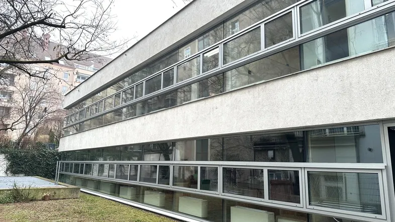 Modernes Gebäude mit vielen Fenstern und einer schlichten Fassade, umgeben von Bäumen und Grünflächen.