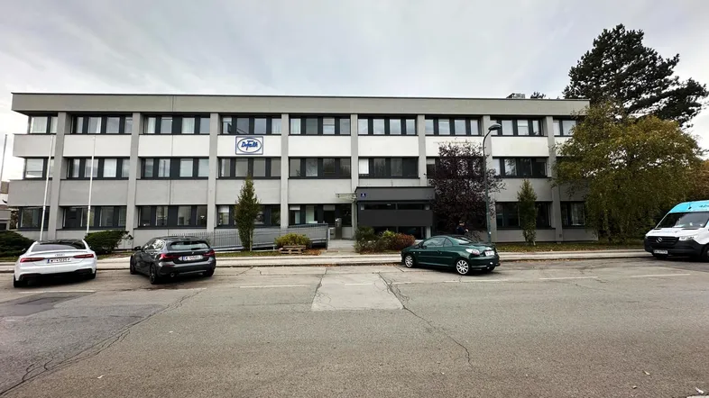 Modernes Bürogebäude mit mehreren Etagen, großen Fenstern und Parkplätzen davor unter bewölktem Himmel.