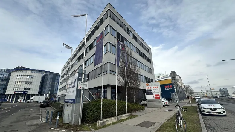 Modernes Bürogebäude mit mehreren Etagen und großen Fenstern an einer belebten Straße unter bewölktem Himmel.