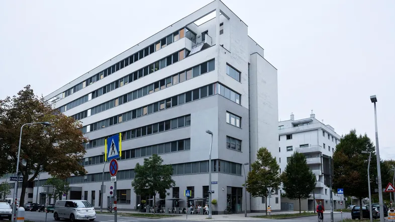 Modernes Bürogebäude an einer Straßenecke mit mehreren Etagen und vielen Fenstern unter bewölktem Himmel.