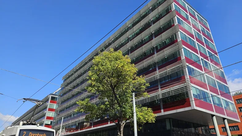 Modernes Bürogebäude mit markanter roter Fassade und großen Glasflächen, im Vordergrund eine Straßenbahn.