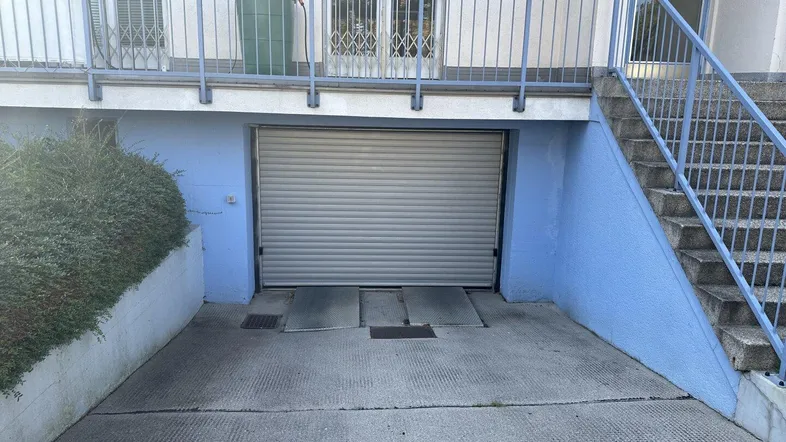 Eingangsbereich zu einer Tiefgarage mit geschlossenem Rolltor und Außentreppe, umgeben von einer Hecke und Blumenkästen.