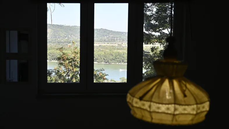 Dunkler Innenraum mit Fensterblick auf einen Fluss und bewaldete Hügel in der Ferne.