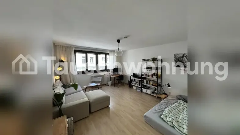 Helles Wohnzimmer mit Holzboden, Sofa, Bett und einem Arbeitsbereich am Fenster.