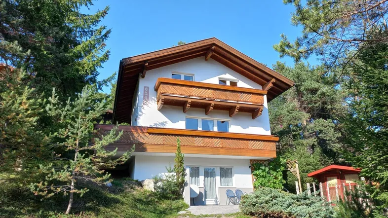 Traditionelles Haus mit Holzbalkonen und Satteldach, umgeben von Nadelbäumen in sonniger Lage.