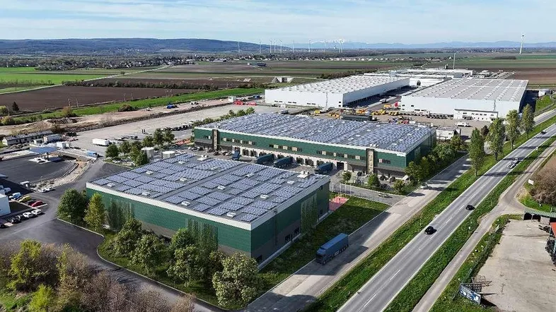 Luftaufnahme eines großen Logistikzentrums mit mehreren Lagerhallen, umgeben von Feldern und einer Straße. Solarpaneele sind auf den Dächern sichtbar.