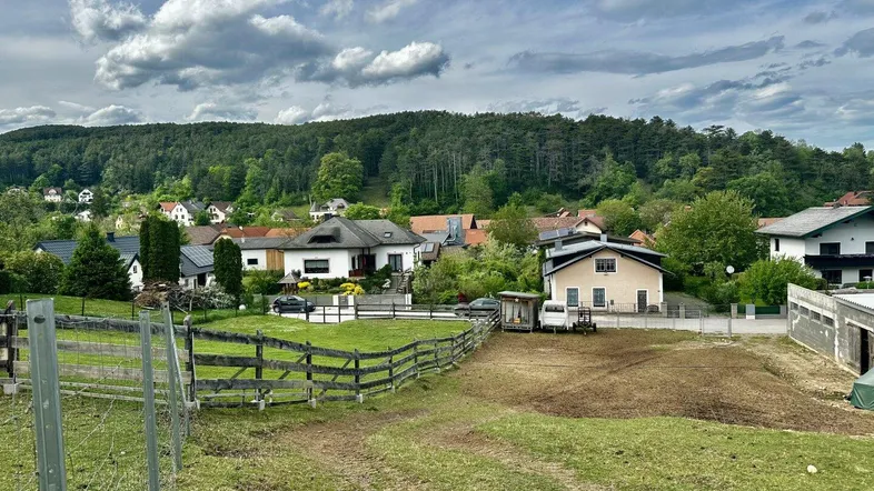 Panoramablick auf eine ländliche Siedlung mit Häusern, grünen Hügeln und Feldern unter bewölktem Himmel.