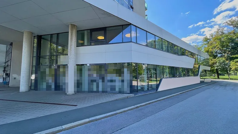 Moderne Gebäudefassade mit großen Glasflächen und klaren Linien, umgeben von Grünflächen und Bäumen.