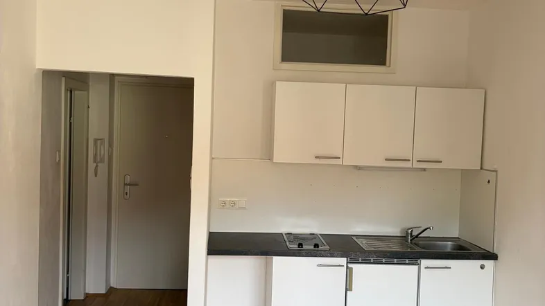 Kleine Küche mit weißen Schränken, Kochfeld und Spüle in einem hellen Raum mit Holzfußboden.