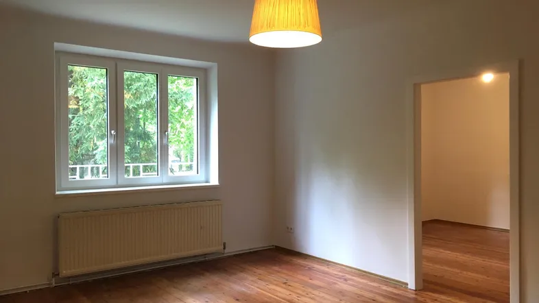 Helles, leeres Zimmer mit Holzdielenboden und Blick ins Grüne durch das Fenster.