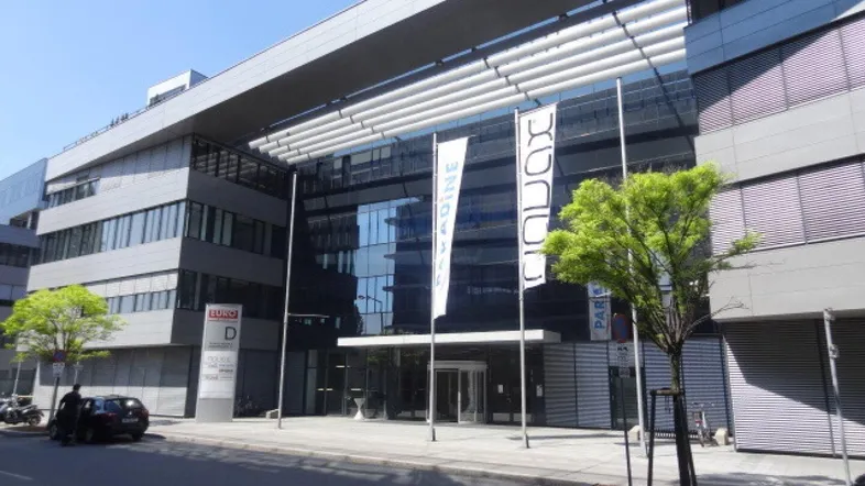 Modernes Bürogebäude mit Glasfassade und breiter Zufahrt, umgeben von Grünflächen.