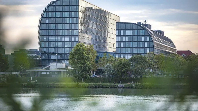 Modernes Bürogebäude mit geschwungener Glasfassade am Flussufer unter bewölktem Himmel.