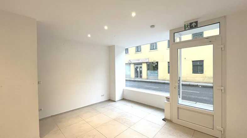 Heller, leerer Geschäftsraum im Erdgeschoss mit großen Fenstern zur Straße und Fliesenboden.