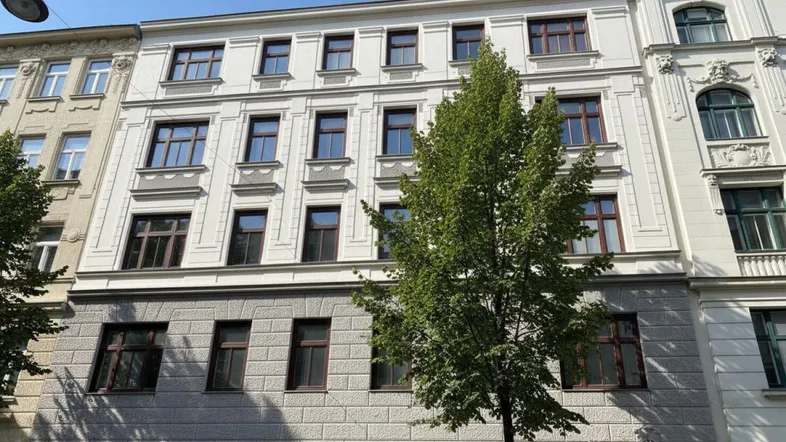Historisches Gebäude mit klassischer Fassade und vielen Fenstern, umgeben von Bäumen und Sonnenschein.