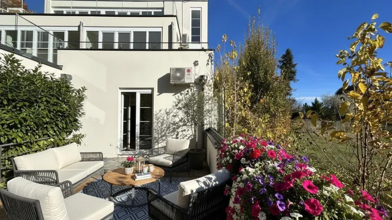 Möblierte Terrasse mit bunten Blumen und Blick auf das Gebäude unter blauem Himmel.