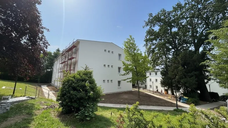 Neubaugebäude mit weißer Fassade und Gerüst, umgeben von viel Grün unter strahlendem Himmel.