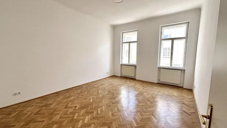 Helles, leeres Zimmer mit Fischgrätparkettboden und zwei hohen Fenstern.