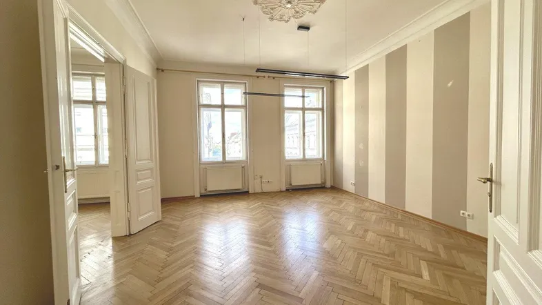 Geräumiges Wohnzimmer mit Fischgrätparkett, hohen Decken und Stuckverzierung, durch große Fenster hell belichtet.