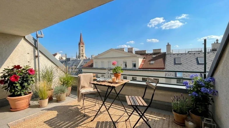 Sonniger Balkon mit Sitzgelegenheiten, Pflanzen und Blick über die Dächer der Stadt mit Kirchturm.