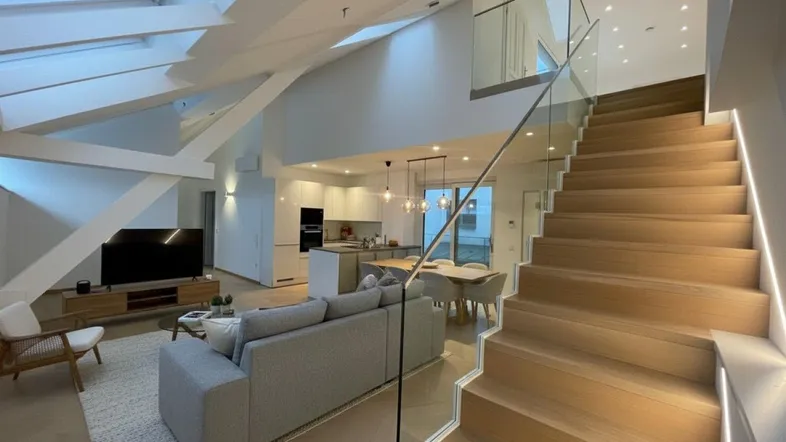 Modernes, offenes Wohnzimmer mit Küche, Essbereich und Holztreppe mit Glasgeländer.