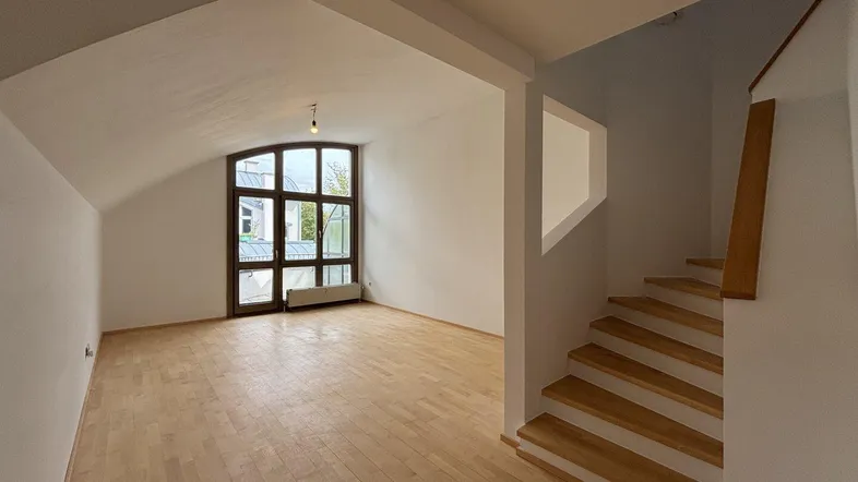 Helles Zimmer mit Holzfußboden, Dachschräge und großem Fenster, das viel Tageslicht hereinlässt.