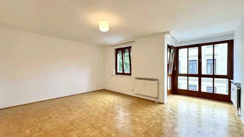 Helles Wohnzimmer mit Parkettboden, weißen Wänden und großen Fenstern, die viel Licht hereinlassen.