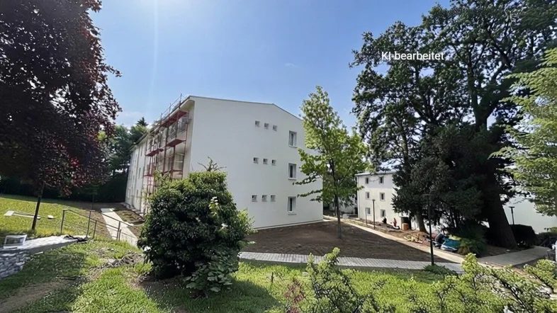 Neubaugebäude mit weißer Fassade und Gerüst, umgeben von viel Grün und Bäumen unter blauem Himmel.