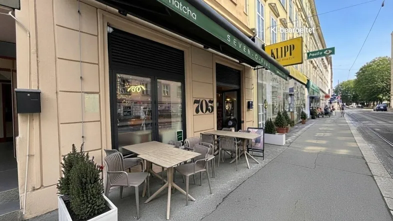 Blick auf die Fassade eines Cafés mit grüner Markise und Tischen im Freien an einem sonnigen Tag.