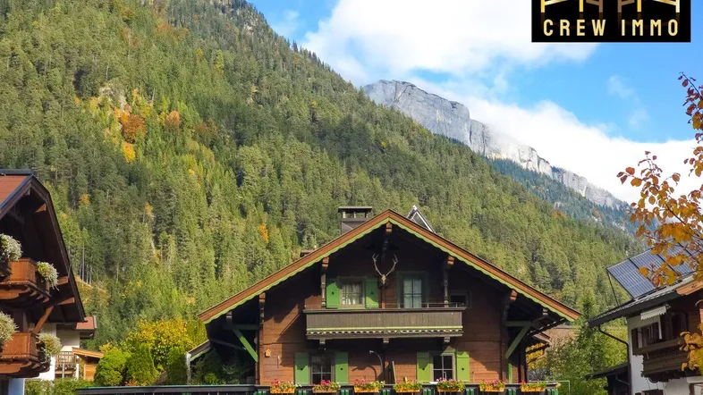 Traditionelles Holzhaus im alpinen Stil mit grünen Fensterläden vor einer bewaldeten Bergkulisse.