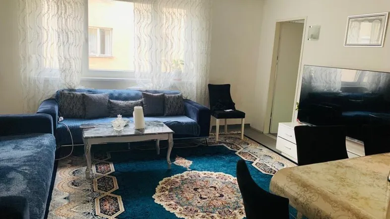 Gemütliches Wohnzimmer mit blauem Sofa, großem Teppich und einem Fenster für natürliches Licht.