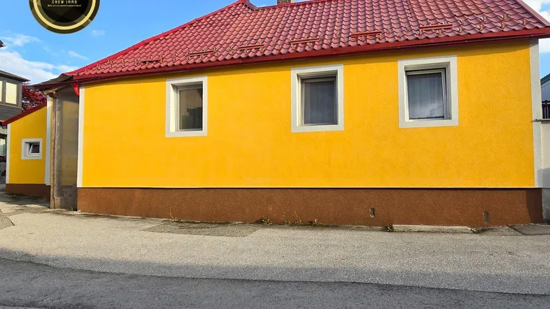 Gelbe Fassade eines Hauses mit rotem Ziegeldach und weißen Fenstern unter blauem Himmel.