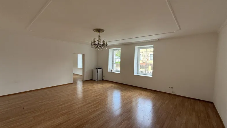 Geräumiges Wohnzimmer mit hellem Laminatboden, hohen Decken und einem eleganten Kronleuchter.