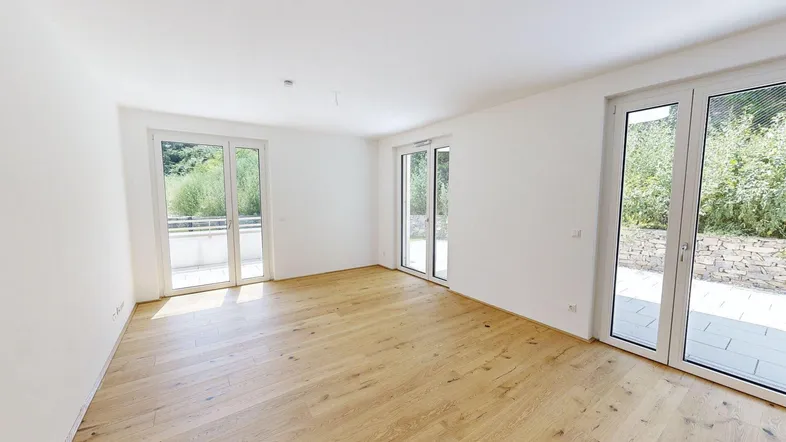 Helles, leeres Zimmer mit Holzboden und großen Fenstertüren, die Zugang zur Terrasse bieten.