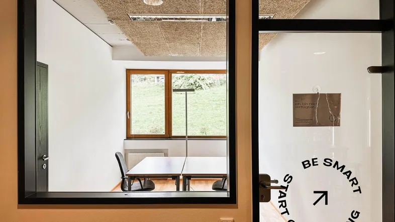 Blick durch eine Glaswand in ein helles Büro mit Schreibtisch, Stühlen und Blick ins Grüne.