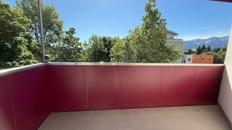 Geräumiger Balkon mit roter Brüstung, Fliesenboden und weitem Blick ins Grüne und auf die Berge.