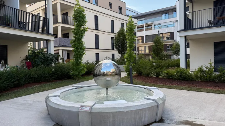 Moderner Brunnen mit spiegelnder Kugel in einer gepflegten Außenanlage zwischen Wohngebäuden.